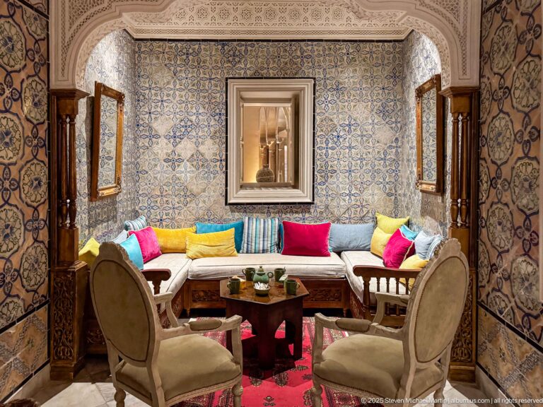 Salon with blue patterned tile walls, cushioned banquette, colorful pillows, mirror, wooden screens, armchairs, and central table.