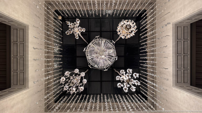 Upward view of several crystal chandeliers and long strands of glass drops suspended inside a tall square lightwell.