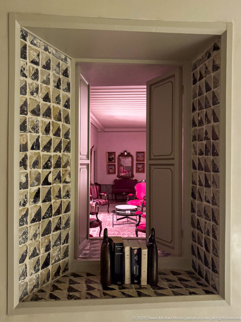 Geometric black-and-cream wall opening with two dark vessels and books, framing a pink-lit sitting room beyond.