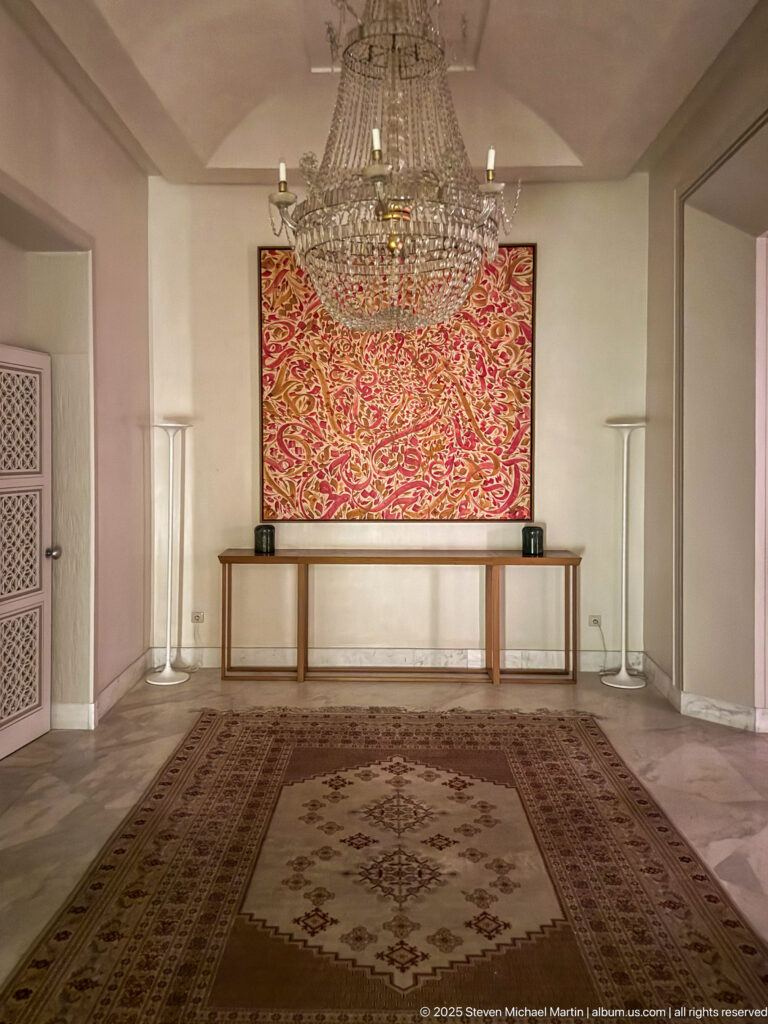 Formal room with large carpet, crystal chandelier, console table, tall lamps, and vivid red patterned artwork on the far wall.