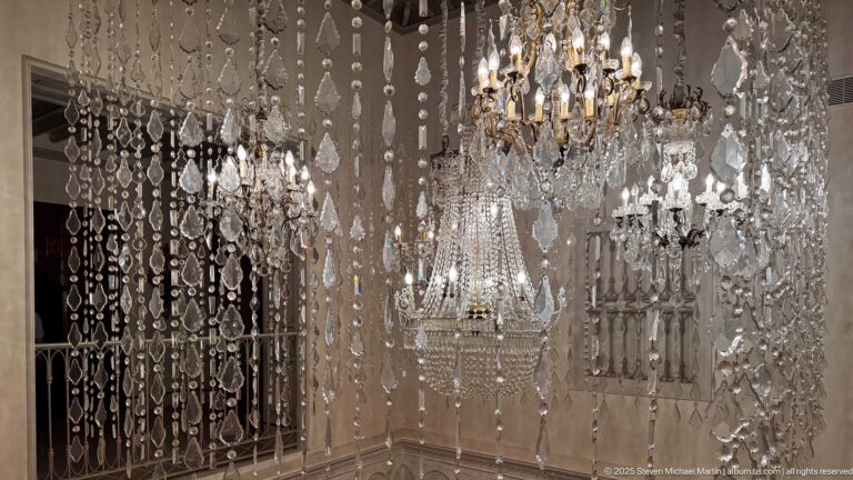 Close view through hanging crystal strands toward several ornate illuminated chandeliers with numerous glass pendants.