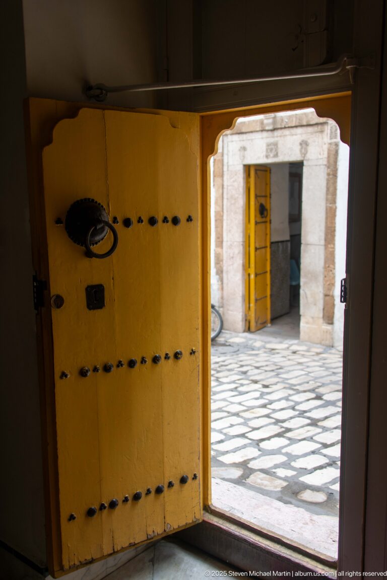 Open yellow timber door with rows of dark metal studs and ring pull, looking into a patterned stone courtyard.