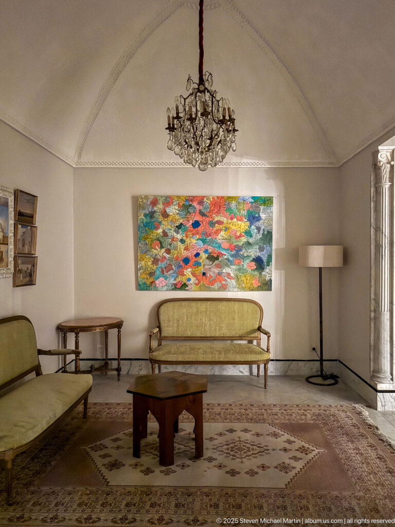 Formal sitting room with green sofas, patterned carpet, wooden tables, dark chandelier, and colorful abstract painting.