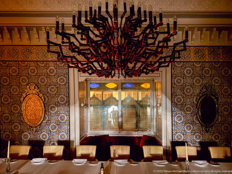 Dining room with patterned blue tile walls, long table, cream chairs, oval wall panels, and a branching red-and-black chandelier.