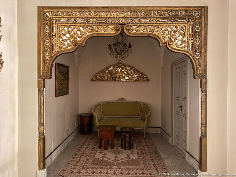 Elaborately carved gilded arch frames a small sitting area with green settee, carpet, tables, and wall decoration.