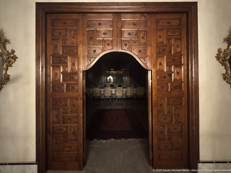 Large dark wooden paneled doorway with an arched opening leading into a dim dining room with pale chairs.
