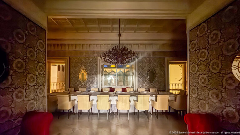 Long dining table with cream chairs beneath a branching chandelier, framed by patterned walls and red velvet seating.