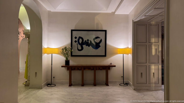 Wooden console beneath black-and-white abstract art, flanked by two yellow-shaded floor lamps and pale paneled doors.