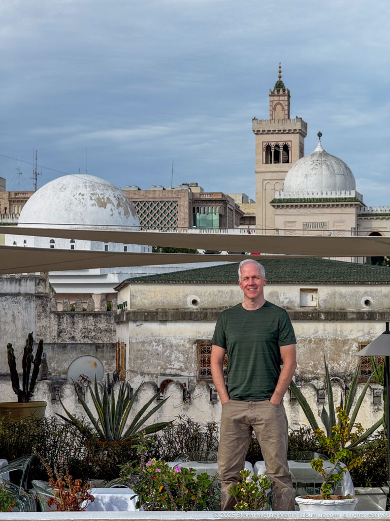 Man in a dark green shirt standing among rooftop plants with white domes and a square tower behind.