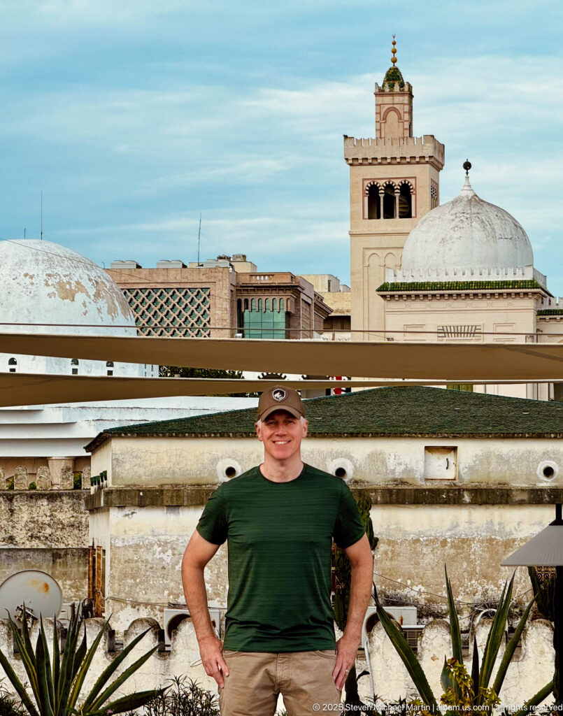 Man in a dark green shirt stands on a rooftop before white domes, a square minaret, latticework, weathered roofs, and plants.