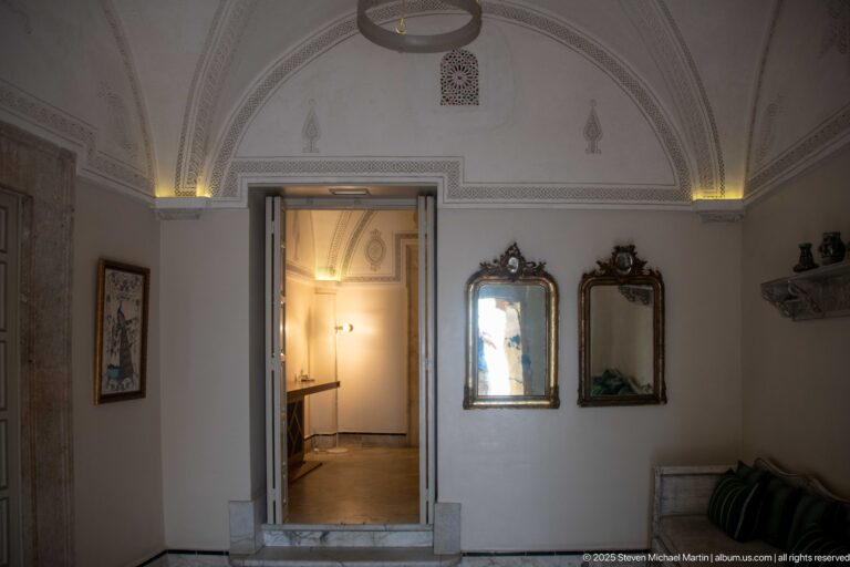 Pale vaulted corridor with two gilded mirrors, a stepped doorway, subtle plaster relief, and warm indirect lighting.