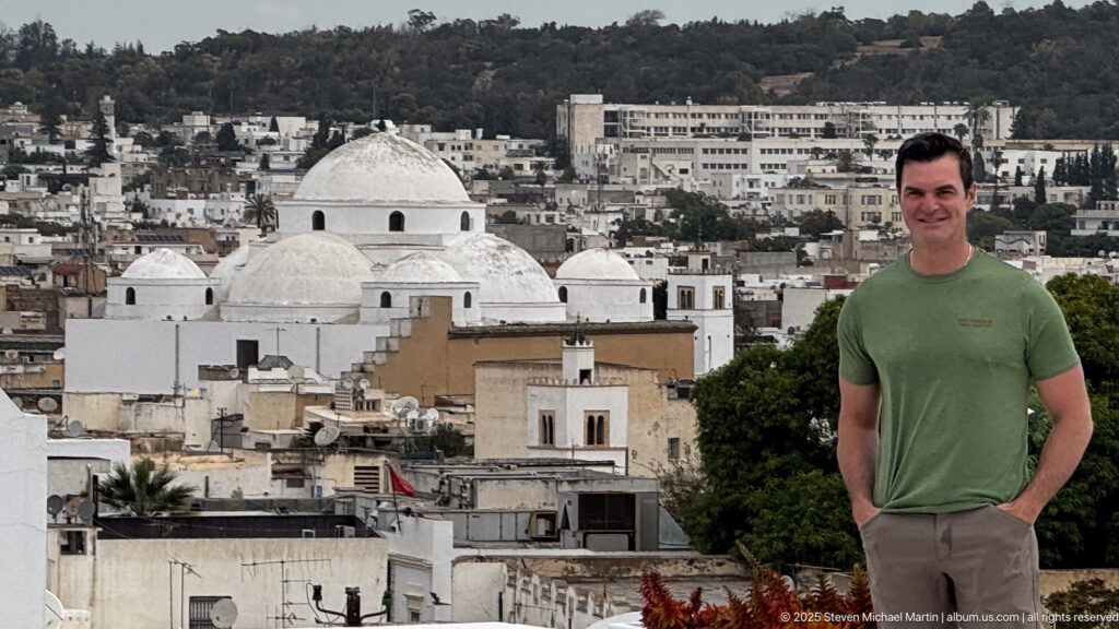 Man in green shirt standing before several white domes, dense Tunis rooftops, and a wooded hillside in the distance.