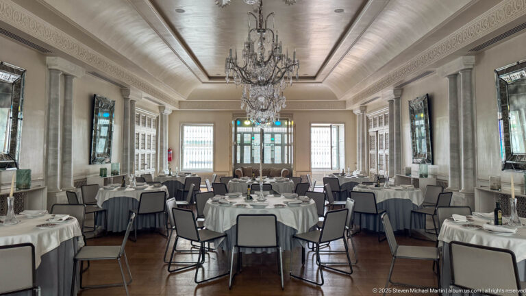 Long formal dining hall with round white-clothed tables, gray chairs, crystal chandeliers, mirrors, columns, and high windows.