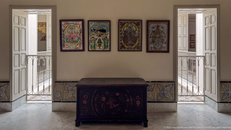 Dark painted chest beneath four colorful framed artworks, between open doorways with white railings and patterned lower wall tiles.
