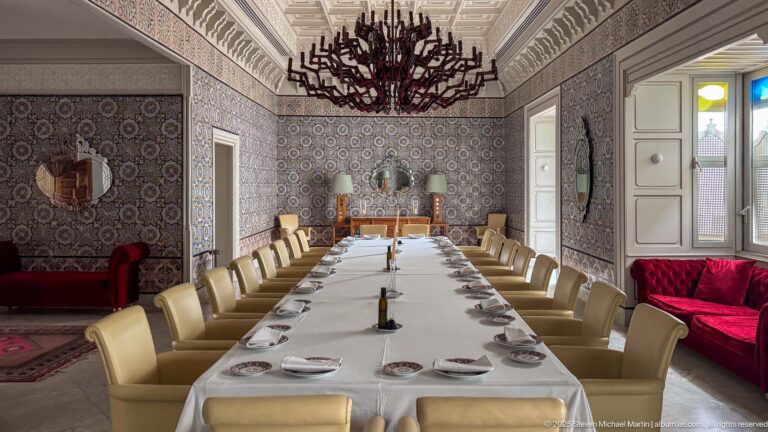 Long white-clothed dining table with cream chairs, patterned tile walls, red velvet seating, and a branching chandelier overhead.