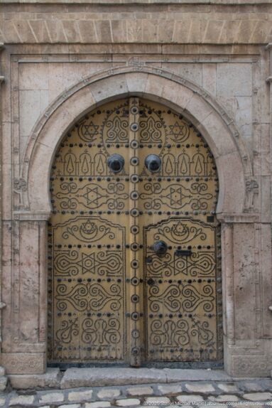 Ornate arched double door with black arabesque patterns and round metal bosses set in a pale stone surround.