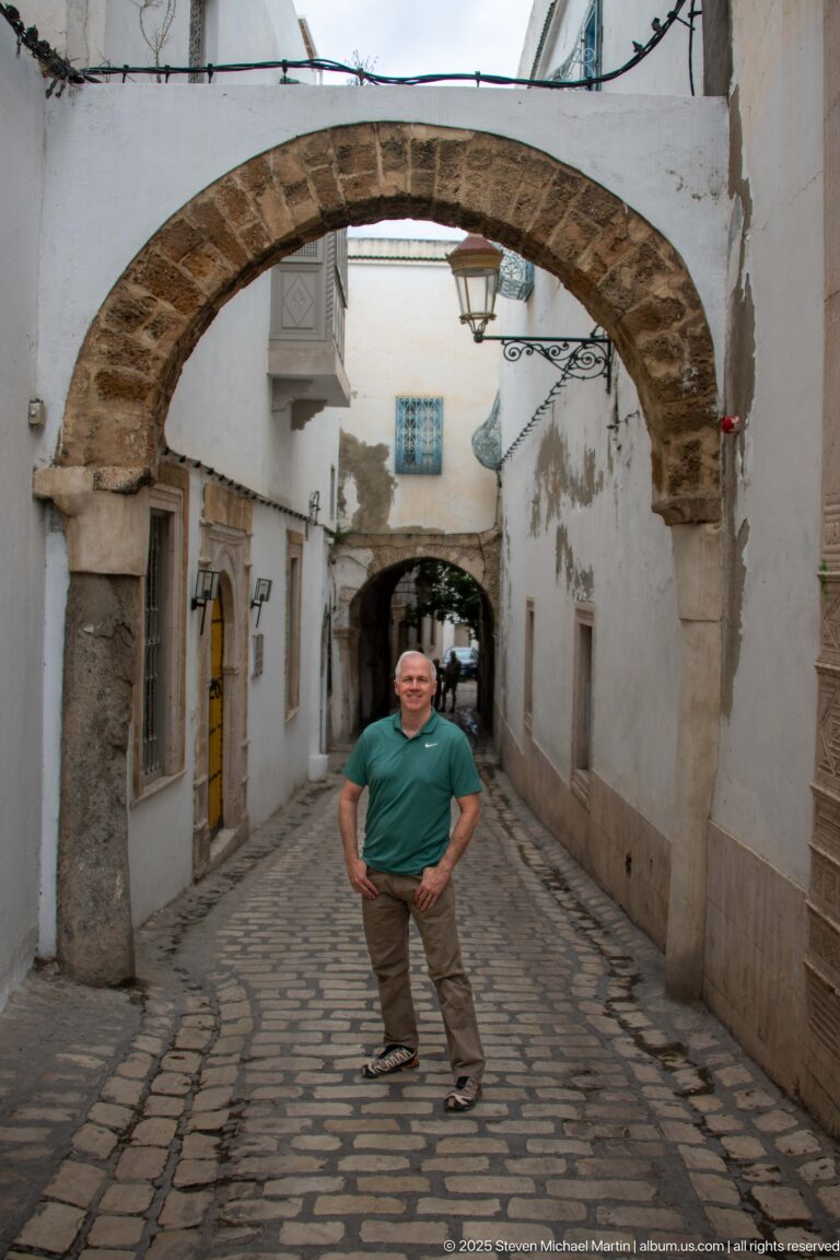 Man standing beneath a broad stone arch on a cobbled medina lane lined with white walls and yellow doors.