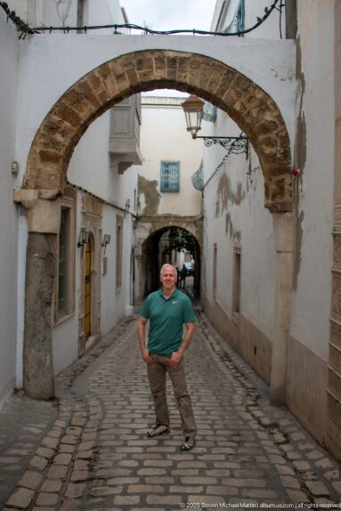 Man standing beneath a broad stone arch on a cobbled medina lane lined with white walls and yellow doors.