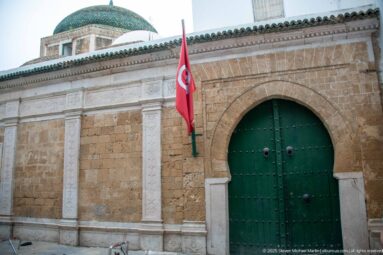 Large green studded arched double door in an ochre stone façade, with a Tunisian flag hanging beside the portal.