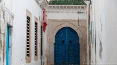 Ornate blue arched double door with dense metal scrollwork and paired knockers at the end of a narrow whitewashed passage.