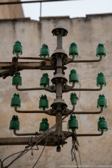 Weathered electrical distribution frame with rows of green ceramic insulators, metal bars, and exposed wiring against a pale wall.