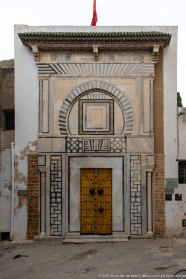 Mustard-yellow studded double door set within an elaborate façade of striped stone arches, geometric panels, pilasters, and tiled cornice.