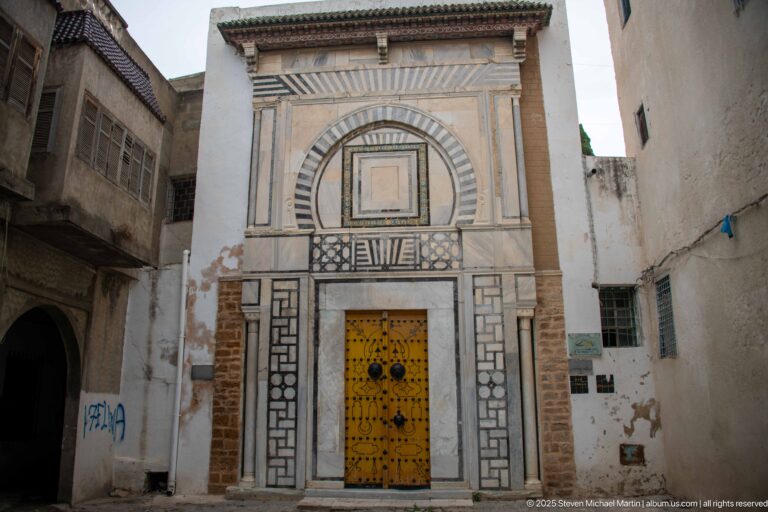 Tall yellow studded double door within an elaborate striped-stone façade of concentric arches and geometric panels on a narrow medina street.