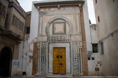 Tall yellow studded double door within an elaborate striped-stone façade of concentric arches and geometric panels on a narrow medina street.