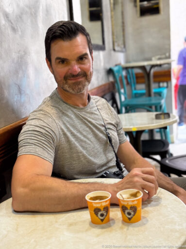 Seated man at a small café table with two orange coffee cups, turquoise chairs, and a softly blurred medina passage behind him.