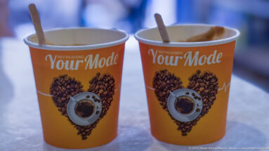 Two orange paper coffee cups with wooden stirrers, decorated with coffee-bean hearts and the words “Refreshing Your Mode.”