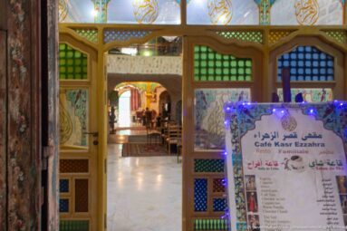 Ornate Café Ksar Ezzahra entrance with colored glass, painted panels, wooden arches, and a multilingual illuminated menu board.