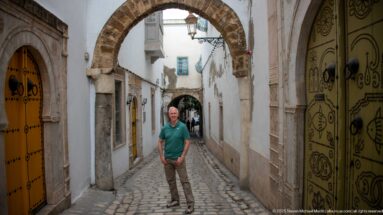 Man standing on a narrow cobbled lane beside ornate yellow doors and beneath a rough stone arch.