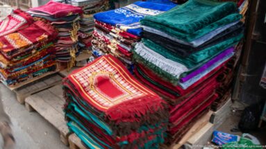 Stacks of folded green, red, blue, pink, and patterned textiles, with a fringed red piece bearing an arched motif on top.
