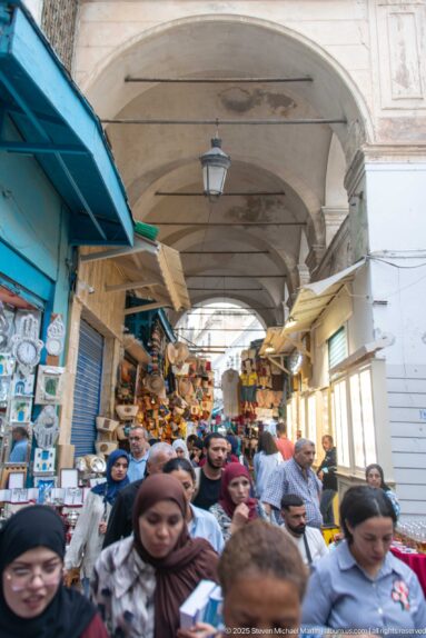 Crowded covered souk passage packed with shoppers beneath shallow vaults, with merchandise and shop displays lining both sides.