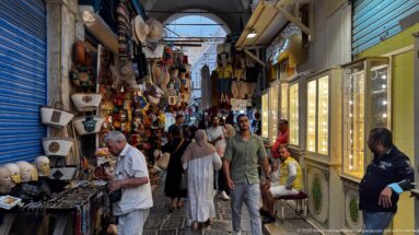 Narrow stone-paved souk lane lined with leather goods, hats, souvenirs, and illuminated display cases, with pedestrians passing between shops.