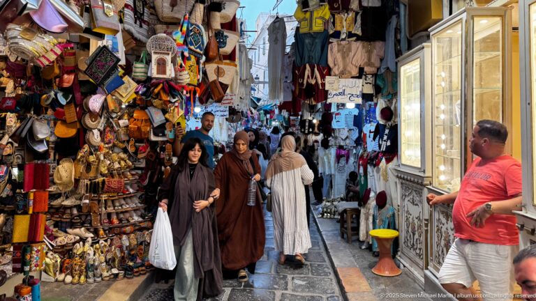 Women walking through a narrow souk lane beside dense displays of leather bags, slippers, hats, souvenirs, and a bright shop case.
