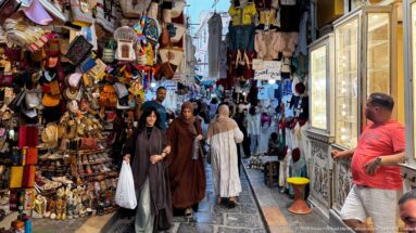 Women walking through a narrow souk lane beside dense displays of leather bags, slippers, hats, souvenirs, and a bright shop case.