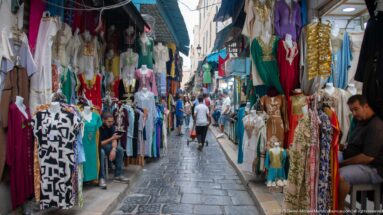 Narrow market lane lined with hanging dresses, tunics, and mannequins, with shoppers walking along the stone-paved center.