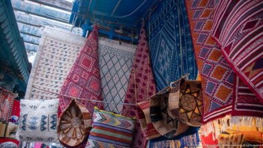 Hanging red, blue, cream, and multicolored rugs above embroidered cushions and leather poufs in a textile market display.