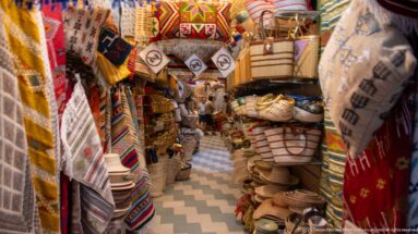 Narrow handicraft souk aisle packed with colorful textiles, woven bags, baskets, hats, and leather goods above a zigzag-patterned floor.