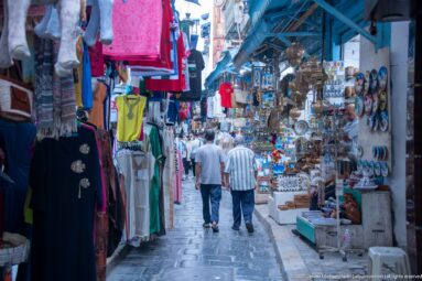 Market lane with hanging clothing at left, metalware and ceramic displays at right, and two men walking away along stone paving.