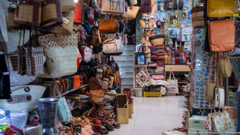Narrow shop interior packed with leather bags, woven baskets, slippers, belts, and small accessories, with stairs rising at the back.