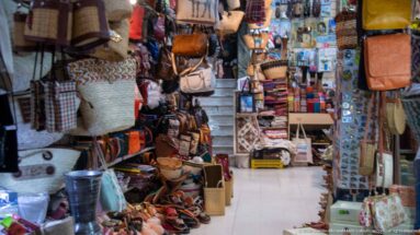 Narrow shop interior packed with leather bags, woven baskets, slippers, belts, and small accessories, with stairs rising at the back.