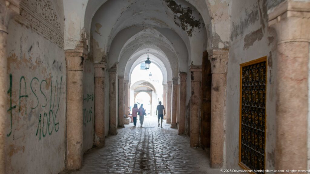 Long vaulted medina passage with stone columns, worn plaster, cobbled paving, and backlit pedestrians near the far opening.