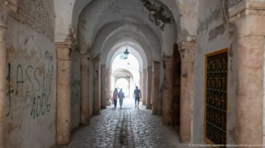 Long vaulted medina passage with stone columns, worn plaster, cobbled paving, and backlit pedestrians near the far opening.