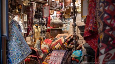 Handicraft shop filled with patterned rugs, cushions, metal lamps, pottery, framed objects, and carved decorative pieces under warm lighting.