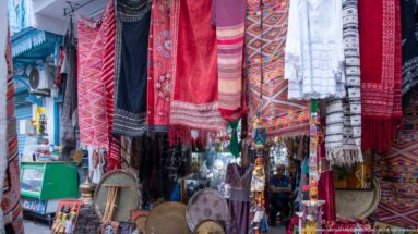 Hanging red, black, white, and multicolored scarves and woven textiles above baskets, drums, and small market goods.