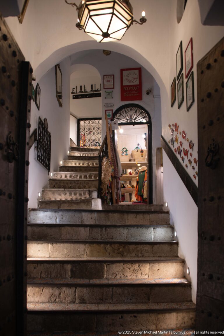 Wide stone staircase with small step lights ascending through a white arch into a decorated interior, flanked by heavy wooden doors.