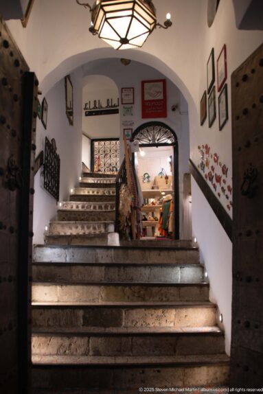Wide stone staircase with small step lights ascending through a white arch into a decorated interior, flanked by heavy wooden doors.