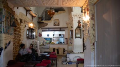 Narrow café interior with exposed stone, red-cushioned stools, hanging lamps, wall niches, mirrors, and two seated patrons.